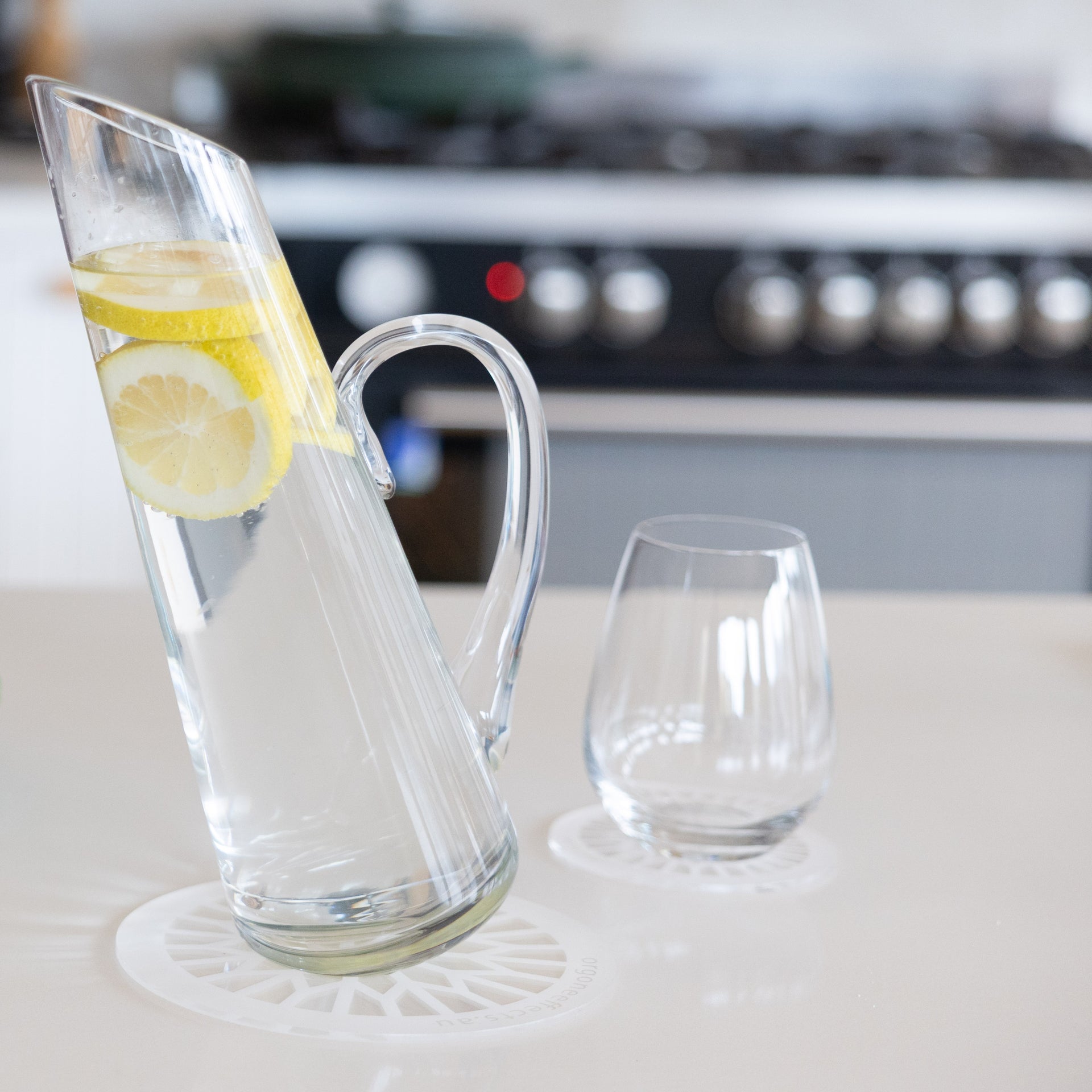 A clear glass pitcher filled with water and lemon slices sits on a white rejuvenation plate next to a matching clear glass tumbler on a kitchen counter, with a blurred stove in the background.