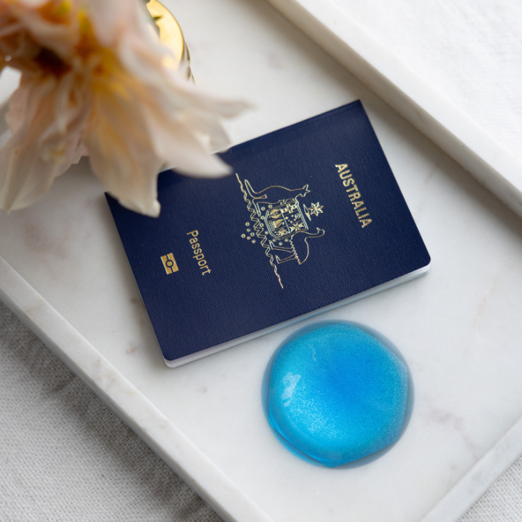 A navy blue Australian passport with gold foil details lies on a white marble tray, next to a round translucent blue EMF harmoniser and a pale flower.
